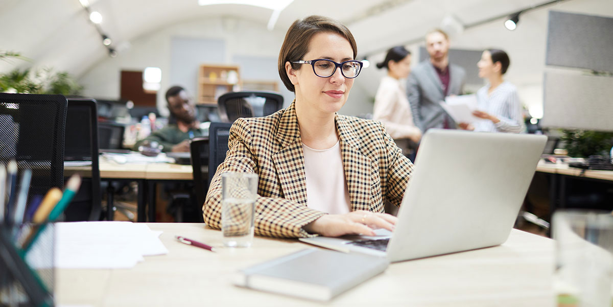 A woman is sitting at a desk, focused on her laptop with colleagues in the background